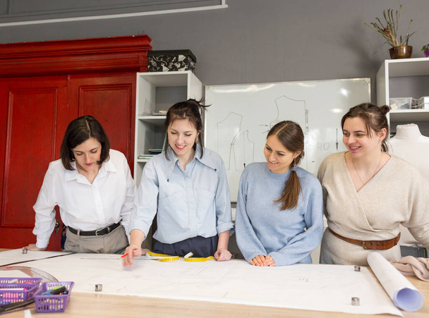 Het concept van kleine bedrijven. Vier vrouwelijke modeontwerpers werken samen in een kleermakersstudio. Een groep damesnaaisters bespreekt stof, patroon en schets in een naaiatelier. - Foto, afbeelding