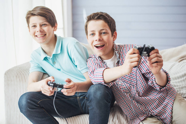 Two attractive teenage boys are playing game console and smiling while sitting on the couch at home - Fotoğraf, Görsel