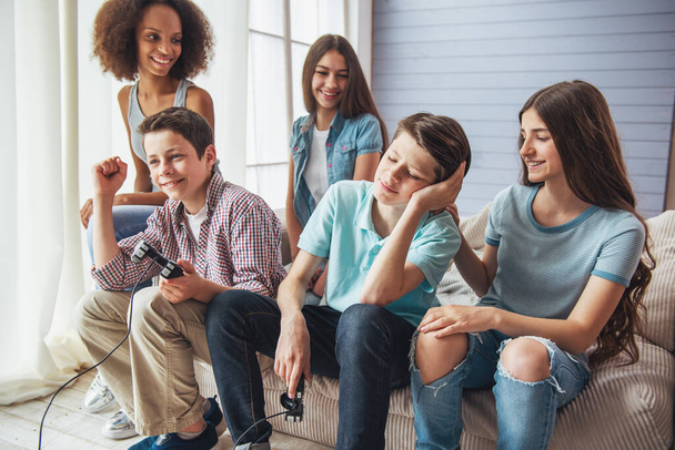 Groupe d'adolescents garçons et filles joue console de jeu et souriant tout en étant assis sur le canapé à la maison
 - Photo, image