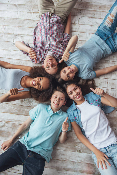 Вид сверху группы мальчиков и девочек-подростков, которые смотрят на камеру, показывают знак ОК и улыбаются, лежа на деревянном полу
 - Фото, изображение