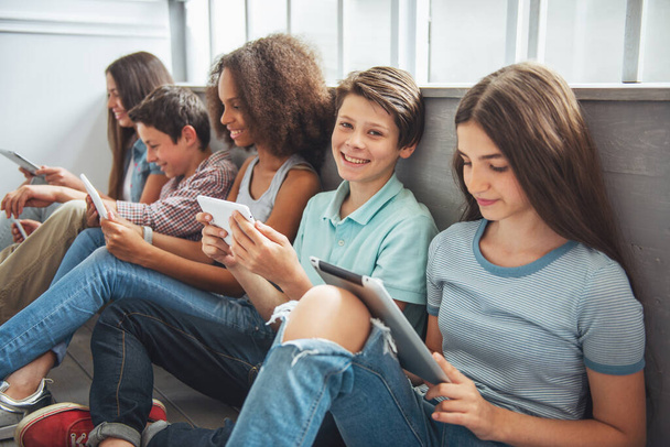 Groep van tiener jongens en meisjes is gebruik van tabletten en glimlachend zittend op de vloer. Een jongen is op zoek op camera - Foto, afbeelding