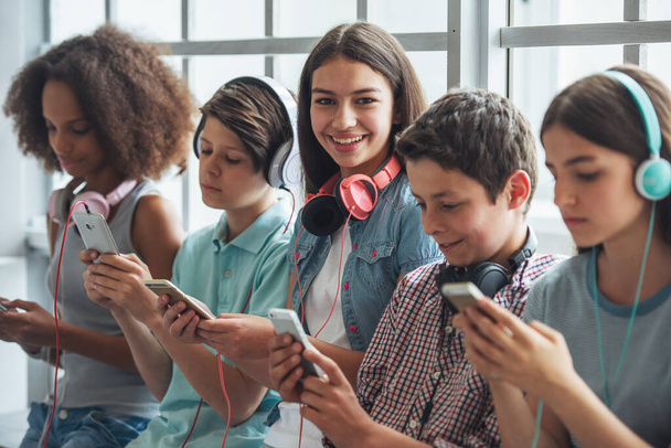 Atrakcyjną nastoletnią dziewczyną z słuchawki jest za pomocą smartfona, patrząc na kamery i uśmiechając się siedząc wśród innych nastolatków - Zdjęcie, obraz