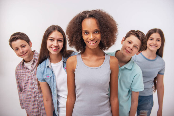 Menina afro-americana muito adolescente está olhando para a câmera e sorrindo, de pé entre outros adolescentes, isolado no branco
 - Foto, Imagem
