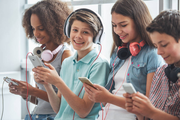 attraktiver Teenager mit Kopfhörern benutzt ein Smartphone, blickt in die Kamera und lächelt, während er zwischen anderen Teenagern sitzt - Foto, Bild