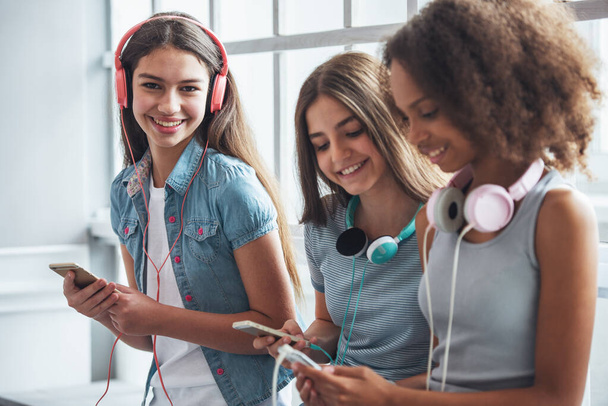 Grupa nastoletnich dziewcząt w słuchawkach jest słuchanie muzyki za pomocą smartfonów i uśmiechając się siedząc przed okno. Jedna dziewczyna jest patrząc na kamery - Zdjęcie, obraz