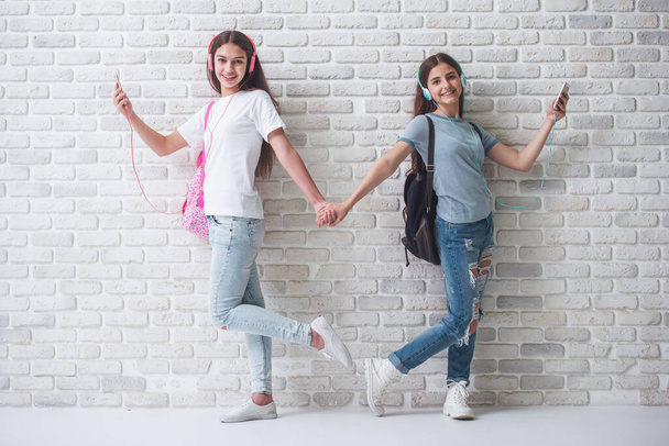Due ragazze adolescenti in cuffia stanno ascoltando musica usando smartphone, tenendosi per mano e sorridendo, in piedi contro il muro di mattoni bianchi
 - Foto, immagini