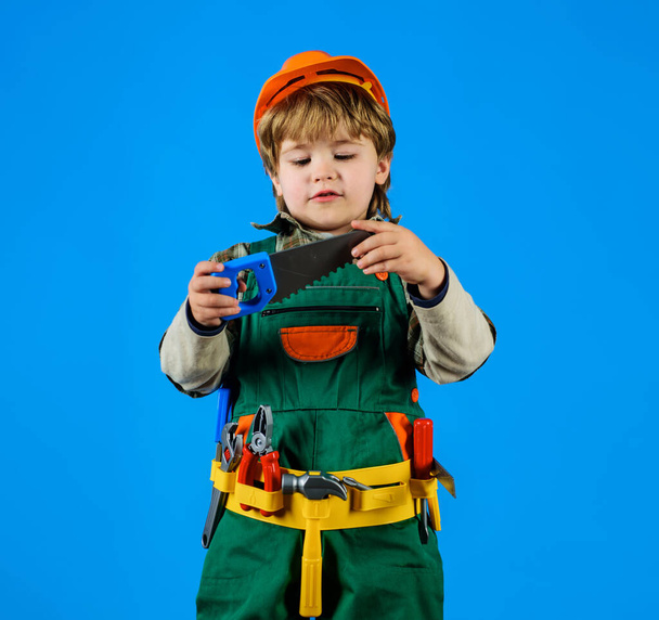 Dítě v bezpečnostní přilbě a opasku s pilou. Pracuj s nářadím. Chlapec v uniformě stavitele s nástrojem na opravu. Malý opravář, řemeslník nebo stavební dělník si hrají s nástroji pro stavbu. Dětská hra - Fotografie, Obrázek