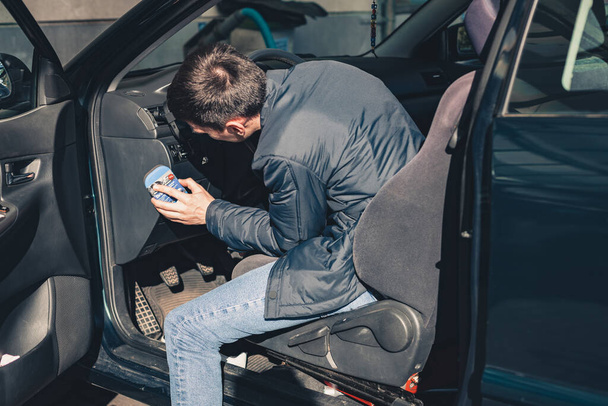 Egy fiatal jóképű fehér férfi letörli a port a kocsi műanyag előlapjáról egy szivaccsal a városi autómosóban egy tiszta tavaszi napsütéses napon, felülnézetből, közelről. - Fotó, kép
