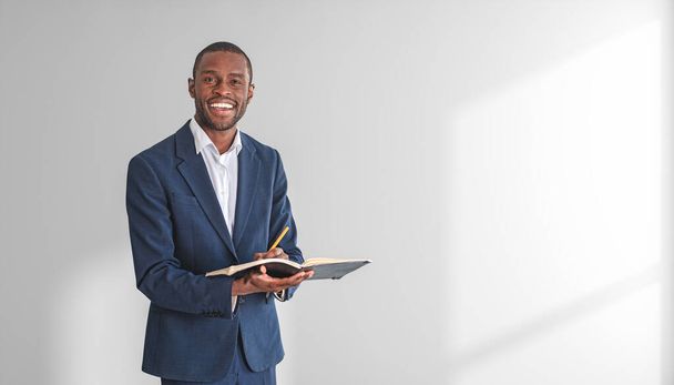 Portrait of cheerful young African American businessman in elegant suit holding notebook and pen and standing near white wall. Concept of planning. Copy space - Fotografie, Obrázek