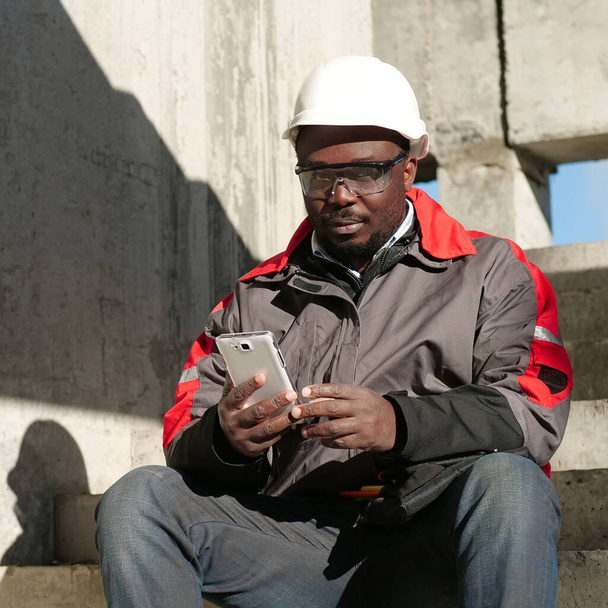 Na staveništi sedí africký americký dělník v klobouku a ochranných brýlích s chytrým telefonem. Dělník drží v ruce bílý smartphone a dívá se do kamery - Fotografie, Obrázek