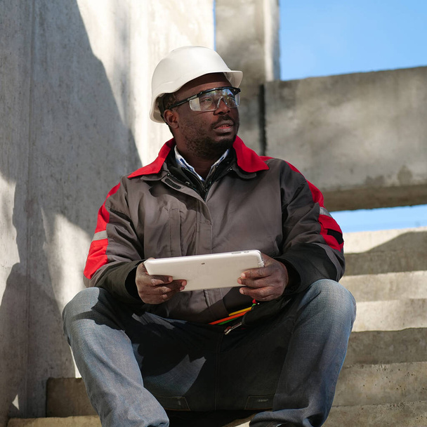 Trabajador afroamericano cansado con los ojos medio cerrados en blanco casco se sienta en el sitio de construcción y sostiene en las manos tableta PC - Foto, imagen