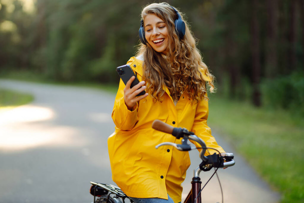 Mulher feliz com fones de ouvido e smartphones monta uma bicicleta em um parque ensolarado ouvindo música. Estilo de vida ativo, férias, descanso. - Foto, Imagem