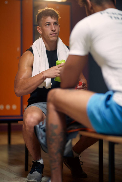 Personal trainer with young man boxer in dressing room, sitting on bench facing each other and talking. Preparation for workout. - 写真・画像