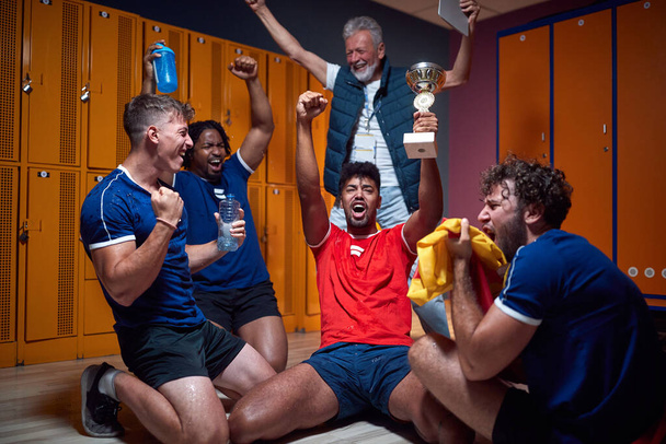Group of young football players celebrating success in the locker room with their senior coach in the background. Sports, active lifestyle concept. - Φωτογραφία, εικόνα