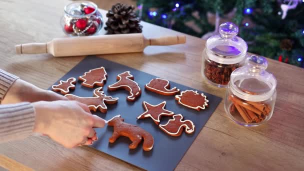 Női kéz színező karácsonyi mézeskalács cookie-kat otthon lassított felvételen. Karácsonyi dekoráció a konyhában. Tündérfényű fenyőfa. Az újév és a karácsony koncepciója. Nyaralás - Felvétel, videó