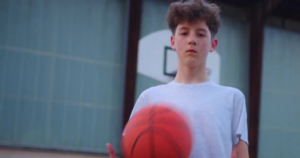 Egy fiatal srác divatos frizurával belenéz a kamerába és egy kosárlabdával játszik. Egy tinédzser portréja a szabadban, iskola után. A modern gyermekek életmódja. - Felvétel, videó