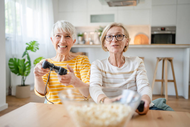 Duas mulheres seniores amigos ou irmãs caucasianas feliz velhos irmãos pensionista jogando console de vídeo game usando joystick ou controladores enquanto sentado em casa pessoas reais família lazer conceito espaço de cópia - Foto, Imagem