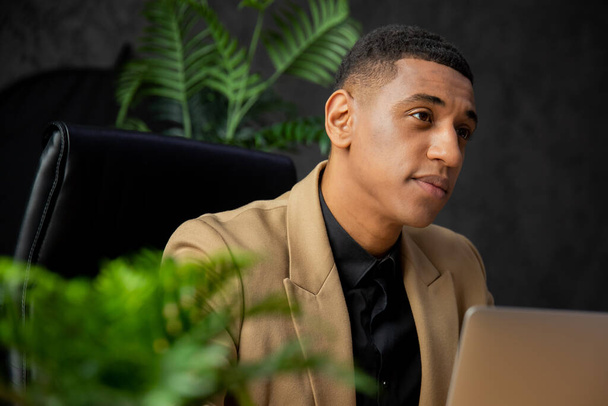 ビジネススーツを着たアフリカ系アメリカ人ビジネスマンが自宅のノートパソコンで働いている。茶色のジャケットを着た若い男がノートパソコンの前の黒い椅子に座っている。高品質の写真 - 写真・画像