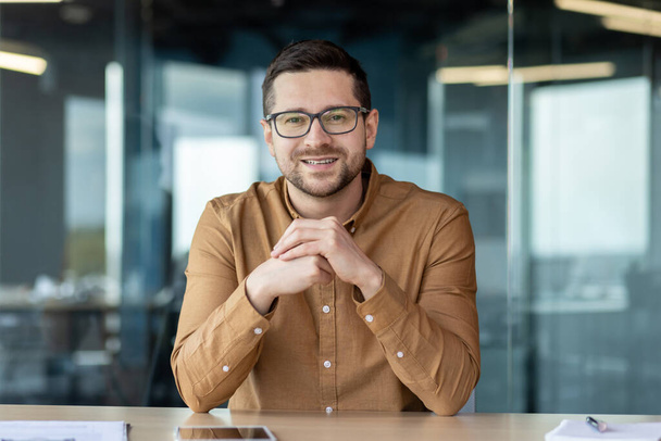 Portrait of a smiling confident man businessman who sits in the office at the table in front of the camera and talks on a video call, conducts business training, consultation. - Foto, afbeelding