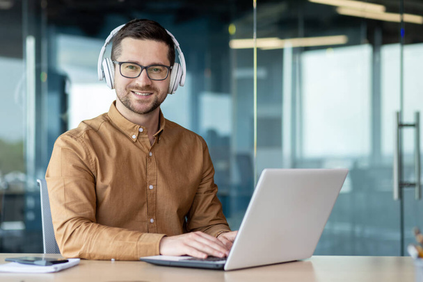 Portrait of a young businessman, a freelancer in headphones who works in an office center at a laptop and looks at the camera with a smile. - Foto, Imagen