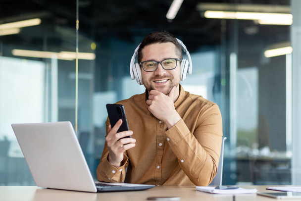Portrait of a young man sitting in the office at the desk wearing headphones, working on a laptop and holding a phone. Smiling and confidently looking at the camera, holding his chin with his hand. - 写真・画像