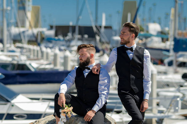 Gay man with partner on wedding day near yacht boat. Married LGBT couples celebrate a romantic wedding ceremony together with a bouquet flower. Portrait of gay couple in love on wedding day - Zdjęcie, obraz