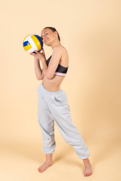 Мила струнка брюнетка в спортивному одязі позує з волейболом. Молодий спортсмен з м'ячем. Ізольовані на пастельному помаранчевому, персиковому фоні
. - Фото, зображення