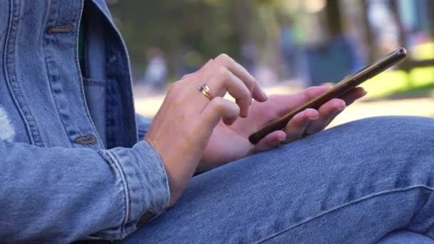 公園に座っているジーンズのデニムジャケットの女性は、彼女のスマートフォンを吸収した。彼女の指は、仮想世界に吸収されたコンテンツをスクロールするときに画面をタップします。自然に囲まれたエンターテイメントは遠くに見えます - 映像、動画