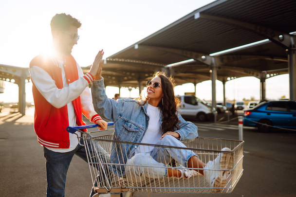Stylish man and woman having fun and riding shopping cart. Black friday. Consumerism, sale, discounts, lifestyle concept. - Fotó, kép