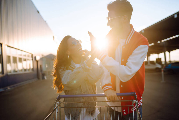 Stylish man and woman having fun and riding shopping cart. Black friday. Consumerism, sale, discounts, lifestyle concept. - Foto, imagen