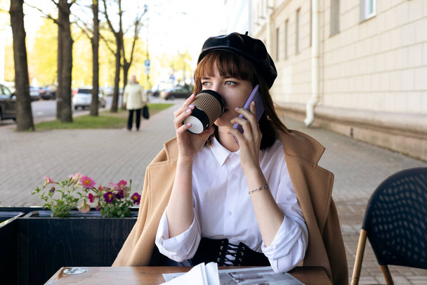 Une belle jeune brune avec une coupe de cheveux bob, portant un béret et un manteau classique beige, est assise sur une terrasse d'un café avec un café, des pâtisseries et un journal, en utilisant le téléphone, appelant. - Photo, image