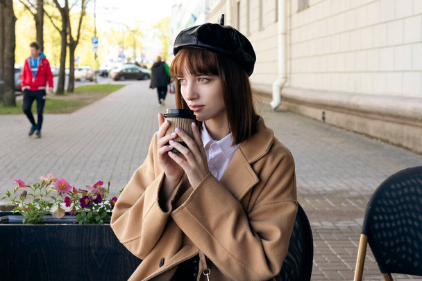 A lovely young brunette with a bob haircut, wearing a beret and a beige classic coat, is sitting on a cafe terrace with a coffee, pastries and a newspaper. Outdoor morning, spring. - Fotoğraf, Görsel