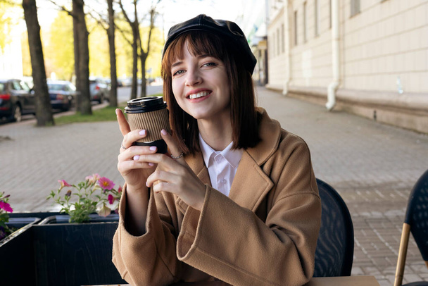 A lovely young brunette with a bob haircut, wearing a beret and a beige classic coat, is sitting on a cafe terrace with a coffee, pastries and a newspaper. Outdoor morning, spring. - Photo, Image