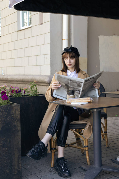 Прекрасна молода брюнетка з боб-стрижкою, одягнена в берет і бежеве класичне пальто, сидить на терасі кафе, читаючи газету
. - Фото, зображення