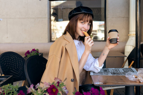 A lovely young brunette with a bob haircut, wearing a beret and a beige classic coat, is sitting on a cafe terrace with a coffee, pastries and a newspaper. Outdoor morning, spring. - Foto, afbeelding