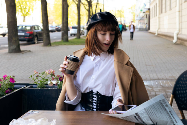 Young brunette with a bob haircut, in a beret and a beige classic coat. Wind blows away a newspaper from a cafe terrace table. Windy day. - Photo, Image