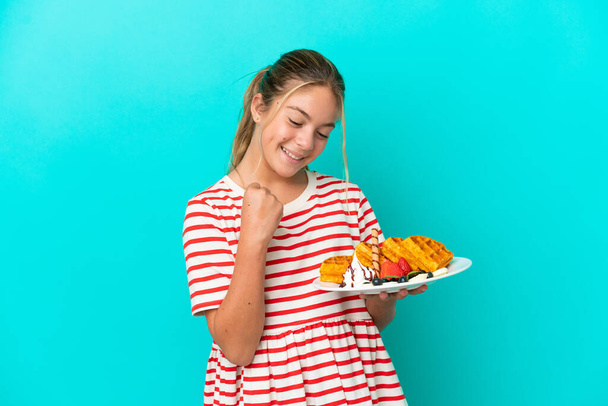 Little caucasian girl holding waffles isolated on blue background celebrating a victory - 写真・画像