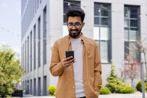Hindu businessman in casual shirt walking near office building from outside, man smiling and using phone, freelancer typing message and browsing internet pages. - Fotografie, Obrázek
