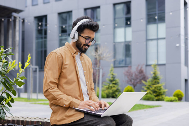 Young successful Hindu student studying online using laptop, man wearing headphones listening to music and online books, audio podcast, typing on keyboard, sitting on bench near university campus. - Фото, изображение