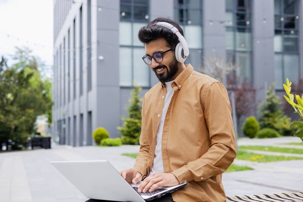 Young successful Hindu student studying online using laptop, man wearing headphones listening to music and online books, audio podcast, typing on keyboard, sitting on bench near university campus. - Foto, Imagem