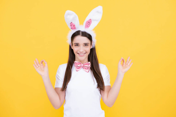 Happy Easter. Portrait of young smiling woman with bunny ears isolated on yellow studio background. Easter bunny woman looks fun. Rabbit girl pointing on copy space - Photo, Image