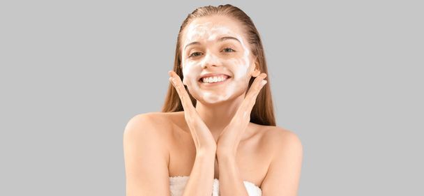 Mooie jonge vrouw met zeepschuim op gezicht tegen grijze achtergrond - Foto, afbeelding