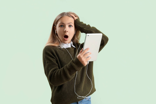 Shocked little girl in earphones with tablet computer on green background - Foto, afbeelding