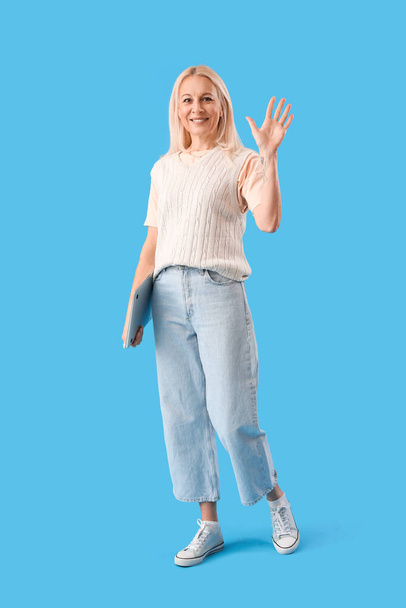 Mature female programmer with laptop waving hand on blue background - Фото, изображение