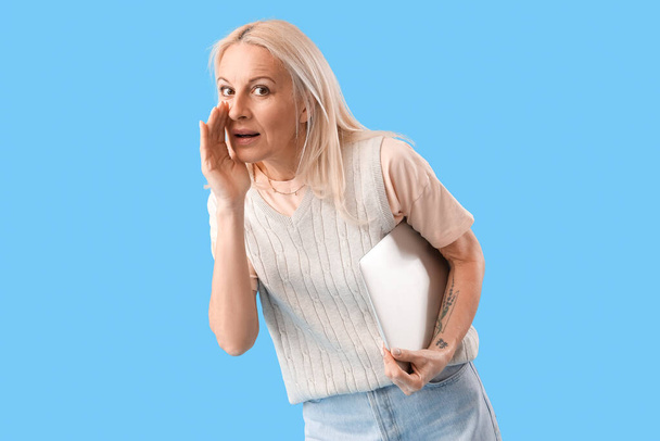Mature female programmer with laptop whispering on blue background - Foto, afbeelding