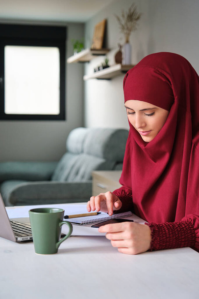 Мусульманская молодая женщина в хиджабе делает ежемесячные счета дома. - Фото, изображение