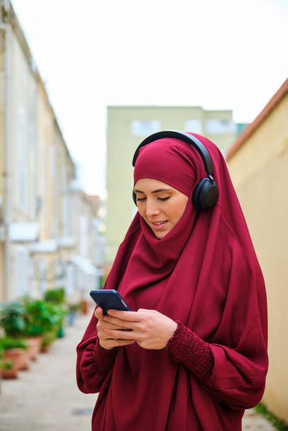 Giovane donna musulmana in hijab utilizzando smartphone e cuffie per strada. - Foto, immagini