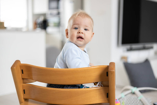 Happy infant sitting in traditional scandinavian designer wooden high chair and laughing out loud in modern bright home. Cute baby smile - Photo, Image
