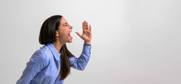 Side view of successful female professional screaming and gesturing in joy while standing isolated on background. Young businesswoman dressed in casuals showing hand sign and shouting - Fotografie, Obrázek