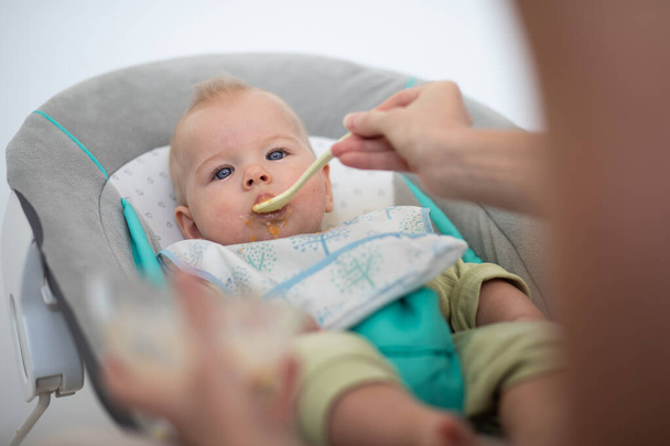 Cuchara madre alimentando a su bebé niño en silla de bebé con puré de frutas. Concepto de introducción de alimentos sólidos para bebés - Foto, Imagen